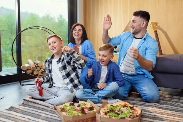 Família elegante feliz sentado no chão, jogando jogos de vídeo com gamepads e comer pizza saborosa. — Fotografia de Stock