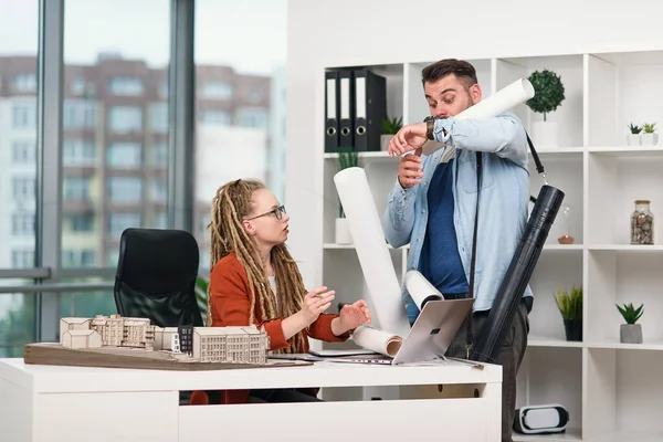 Serious female manager of design agency is upset as clumsy worker let out blueprints on her table.