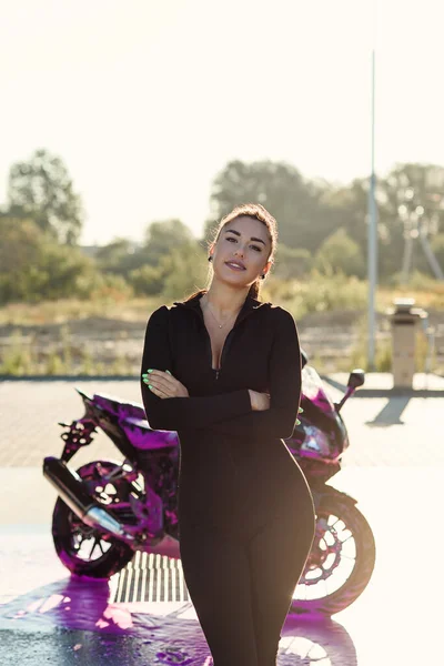 Seductive young woman in tight fitting black suit poses near sport motorcycle at self service car wash. — Stock Photo, Image