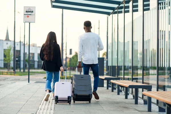 Stylowa dziewczyna i czarny facet niosące walizki na kołach posiadających paszporty z biletami i chodzenie po dworcu autobusowym. — Zdjęcie stockowe