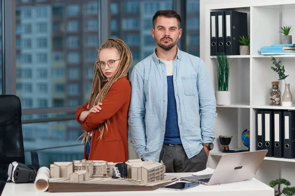Handsome professional young engineer stands back to back with pretty female colleague with dreadlocks at design bureau.