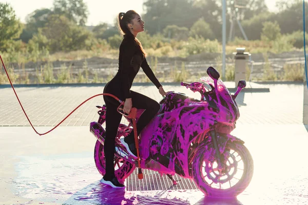 Menina bonita em terno sedutor preto fica perto de moto na lavagem de carro self-service ao nascer do sol. — Fotografia de Stock