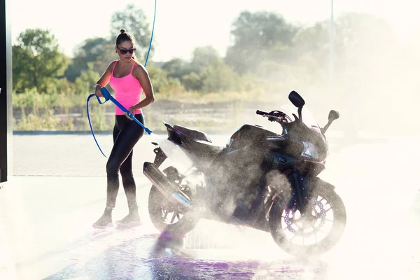Красивая потрясающая девушка моет мотоцикл в автомойке самообслуживания с струей воды высокого давления утром на рассвете. Лицензионные Стоковые Изображения