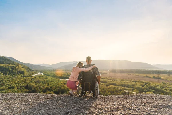 Beautiful woman hugs her disabled husband in wheelchair on a mountain hill at sunset. — Stock Photo, Image