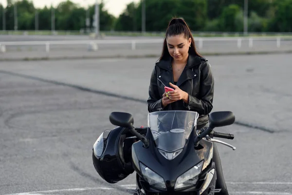 Bonito motociclista feminino usa smartphone enquanto sentado em motocicleta esporte elegante no estacionamento ao ar livre. — Fotografia de Stock