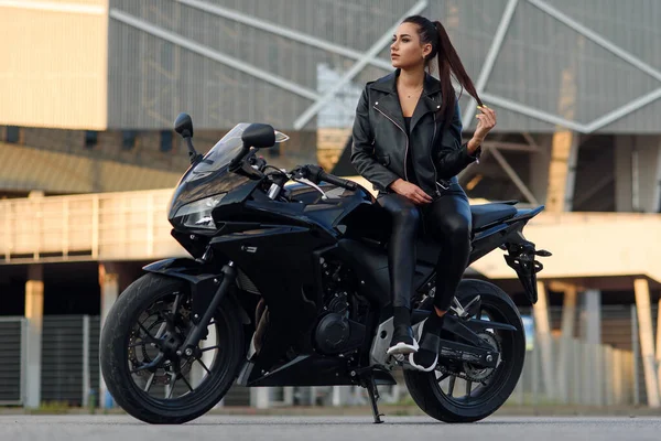 Chica atractiva con el pelo largo en chaqueta de cuero negro y pantalones en el aparcamiento al aire libre con la motocicleta deportiva con estilo al atardecer. — Foto de Stock