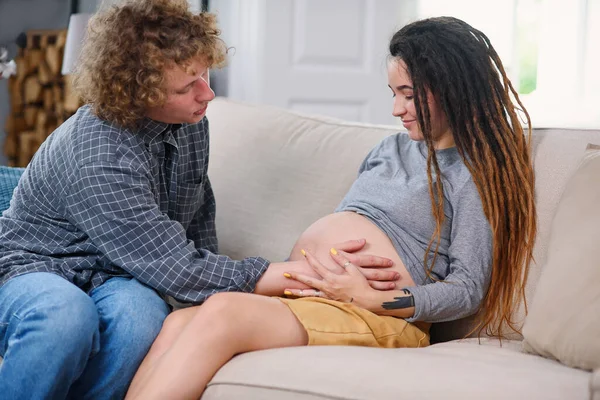 Férj fogdossa terhes has bájos örömteli felesége, amely feküdt a kanapén a hangulatos nappaliban — Stock Fotó