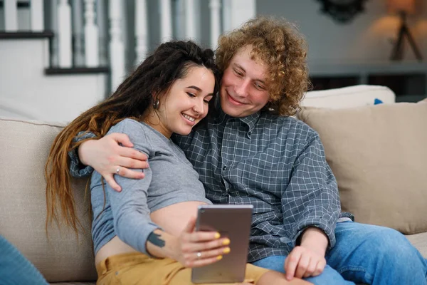 Goed uitziende zwangere paar ontspannen op de comfortabele bank thuis en tv-shows kijken op tablet pc. — Stockfoto