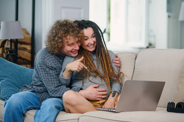Kochający mężczyzna siedzi ze swoją ciężarną żoną ze stylowymi dredami na kanapie i rozmawia wideo na komputerze. — Zdjęcie stockowe