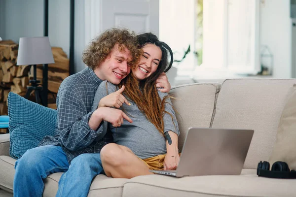 Loving man zit met zijn zwangere vrouw met stijlvolle dreadlocks op de bank en het hebben van video chat op de computer. — Stockfoto