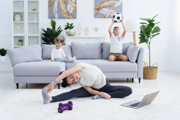 Exercícios de alongamento da mãe enquanto as crianças ativas energéticas brincam no fundo. Mãe fazendo exercício de ioga em casa. — Fotografia de Stock