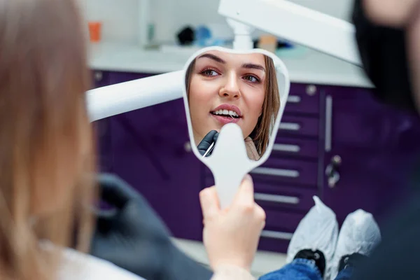 Paciente jovem olhando para o espelho e admira seu novo sorriso após o tratamento odontológico na clínica odontológica. — Fotografia de Stock