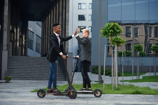 Joyful trabajadores de oficina internacionales que montan en scooters eléctricos y dan choca los cinco en movimiento. — Foto de Stock