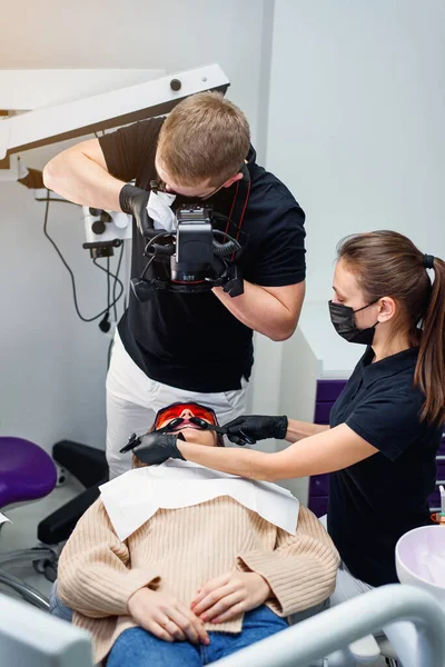 助手付き歯科医は、歯科椅子に座っている女性患者の歯の口腔内写真を撮る. — ストック写真
