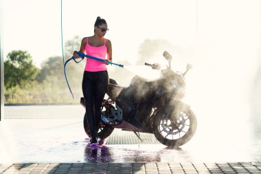 Güzel, çarpıcı bir kız, sabah güneş doğarken yüksek basınçlı su jeti ile oto yıkamada motosikleti yıkıyor..