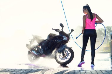 Güzel, çarpıcı bir kız, sabah güneş doğarken yüksek basınçlı su jeti ile oto yıkamada motosikleti yıkıyor..