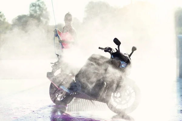 Hermosa chica impresionante lava una motocicleta en auto servicio de lavado de coches con chorro de agua de alta presión en la mañana al amanecer. —  Fotos de Stock