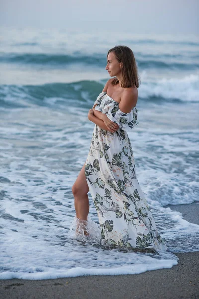 Jovem mulher bronzeada bonita vestindo um vestido elegante posando em pé na praia perto de ondas e olhando para o mar. — Fotografia de Stock