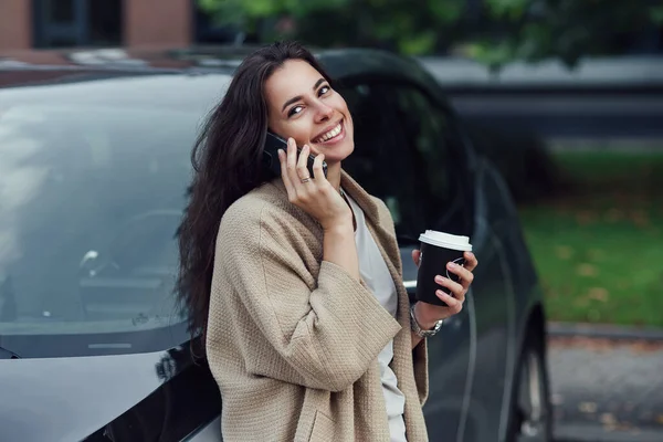 Giovane bella donna sorridente con una tazza di caffè parla su smartphone vicino al suo veicolo elettrico. — Foto Stock
