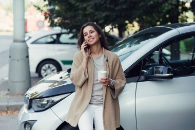Elinde kahve fincanı olan genç ve güzel bir kadın elektrikli aracının yanında akıllı telefondan konuşuyor..