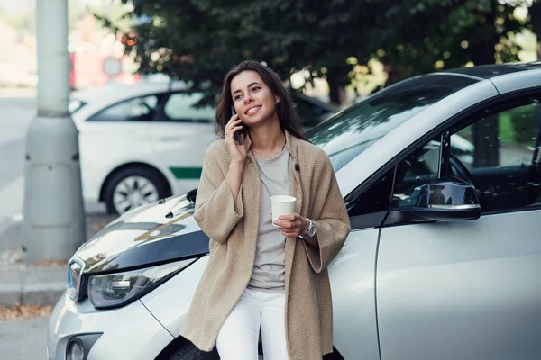 Jovem bela mulher sorridente com xícara de café fala no smartphone perto de seu veículo elétrico. — Fotografia de Stock