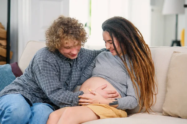 Make röra gravid mage av hans charmiga glada fru som ligger på soffan i mysiga vardagsrum — Stockfoto