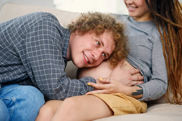 Husband touching pregnant tummy of his charming joyful wife which lying on the couch in cozy living room — Stock Photo, Image