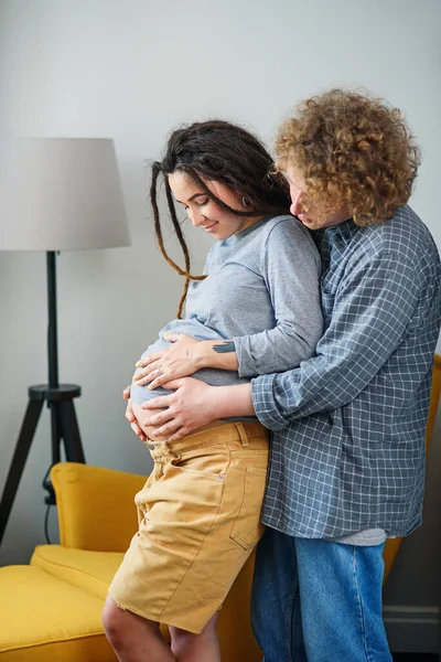 Ευτυχισμένο hipster έγκυος ζευγάρι στέκεται μαζί κοντά στο παράθυρο στο σύγχρονο διαμέρισμα και τρυφερά αγγίζοντας κοιλιά. — Φωτογραφία Αρχείου