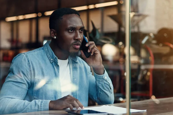 Guapo hombre barbudo de piel oscura con estilo sentado cerca de la ventana de la cafetería de la calle y habla en el teléfono inteligente con el socio de negocios. — Foto de Stock