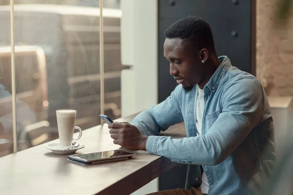 Empresario negro concentrado utiliza teléfono inteligente mientras está sentado en la cafetería con café cerca de la ventana. — Foto de Stock
