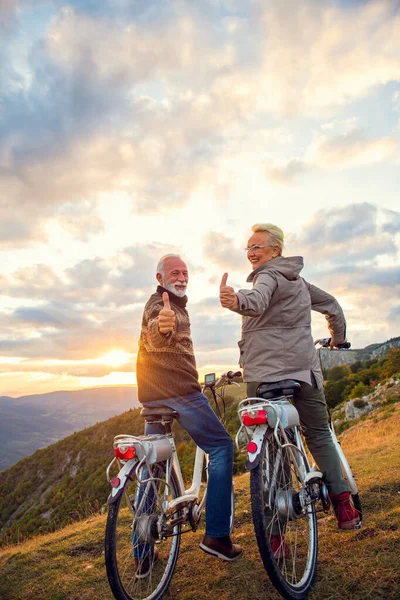 Hombre y mujer mayores en bicicletas eléctricas sosteniendo sus pulgares en la montaña. Hermosa puesta de sol en el fondo. —  Fotos de Stock