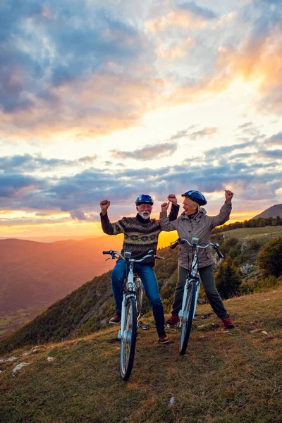 Feliz pareja de ancianos montando bicicletas en el parque y levantando las manos —  Fotos de Stock