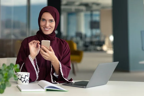 Selamat muslim pengusaha dalam jilbab di kantor tempat kerja. Smiling Arabic woman working on laptop and using smatphone. — Stok Foto