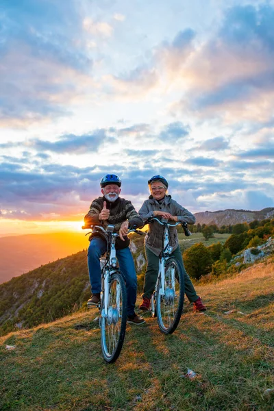 Ηλικιωμένοι άνδρες και γυναίκες με e-bikes κρατώντας τους αντίχειρες τους επάνω στο βουνό. Όμορφο ηλιοβασίλεμα στο φόντο. — Φωτογραφία Αρχείου