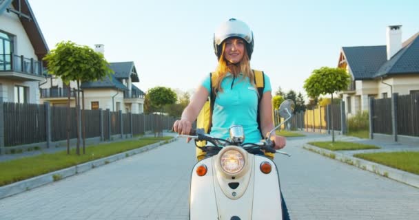 Wesoła biała kurierka w kasku, dostarczająca jedzenie z termalnym plecakiem. Ładna młoda kurierka siedząca na skuterze, patrząca i uśmiechnięta do kamery. Zakupy online, handel elektroniczny. — Wideo stockowe