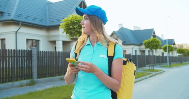 Bellissimo corriere caucasico con uno zaino termico che consegna il cibo. Giovane operaia consegna visualizzazione mappe e alla ricerca dell'indirizzo del cliente e sorridente quando trovare casa. Shopping online. — Video Stock