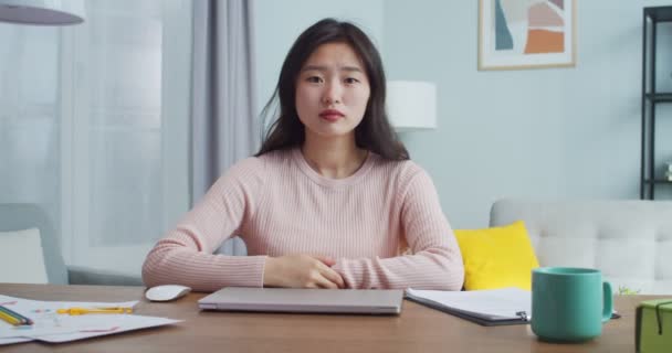 Ładna młoda kobieta w nowoczesnym pokoju biurowym o minimalistycznym wzornictwie. Azjatycka studentka w luźnych ciuchach siedząca przy stole i patrząca w kamerę. Uczenie się na odległość, praca w domu. — Wideo stockowe