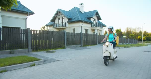 Wanita Kaukasia pekerja pengiriman naik skuter turun ke jalan mencari alamat pelanggan. Gadis kurir dengan helm dan termal ransel memberikan pesanan. E-commerce dan belanja online — Stok Video