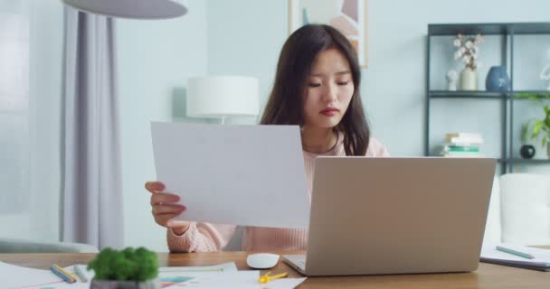 Linda freelancer asiática sentada à mesa, trabalhando em casa. Mulher jovem inteligente segurando folha de papel, digitando no computador portátil, verificando documentos. Negócios, comércio. — Vídeo de Stock