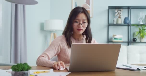 Mulher asiática atraente em roupas casuais sentado à mesa no quarto do escritório. Jovem estudante freelancer em óculos abrindo laptop para trabalhar e estudar em casa distantemente. Interior minimalista moderno. — Vídeo de Stock