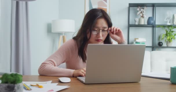 Piękna azjatycka studentka siedzi przy stole i naprawia okulary czytając wiadomości, e-maile w domu. Szczęśliwa studentka, używająca laptopa do nauki. Edukacja, praca, koledż, przyjęcie na uniwersytet. — Wideo stockowe