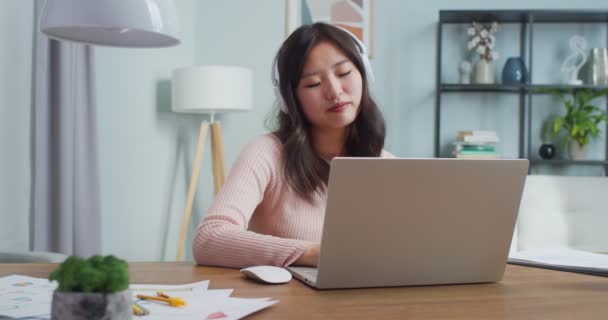 Hübsche Asiatin sitzt am Tisch und arbeitet, studiert im Homeoffice. Entspanntes junges Mädchen mit Kopfhörer, Laptop und Kopfschütteln beim Musikhören. Internet, Fernunterricht. — Stockvideo