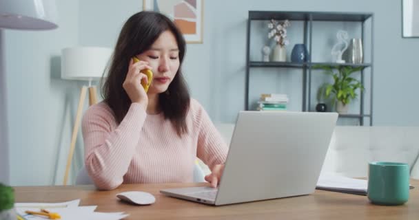 Uma trabalhadora asiática sentada à mesa e a falar ao telefone com o parceiro. Mulher adulta inteligente usando laptop, discutindo projeto com colega, chefe e digitação. Cotonete de negócios. — Vídeo de Stock