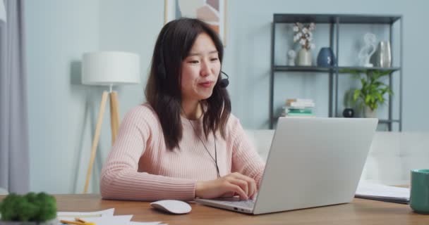Okouzlující Asiatka sedící u stolu a používající notebook v domácí kanceláři. Usmívající se mladá dívka ve sluchátkách mluví s rodinou, přátelé přes webkameru, video chat, gestikulace. — Stock video