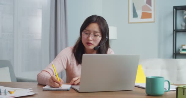 Trabalhador asiático muito sentado à mesa e falando com os clientes, cliente via chat webcam de vídeo. Mulher adulta trabalhadora em fones de ouvido e óculos trabalhando em casa e tomando notas. Serviço de apoio. — Vídeo de Stock