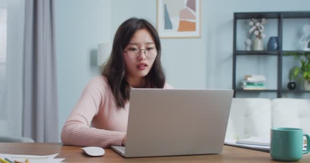 Cansada jovem estudante em glaavalie sentado à mesa e estudando em casa. Mulher asiática exausta terminando o trabalho, tirando óculos e fechando laptop ceen. Educação, ensino à distância. — Vídeo de Stock