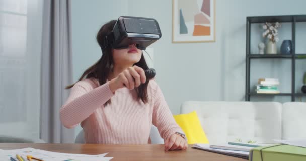 テーブルに座って、ミニマルなデザインのモダンな部屋で楽しんでいる若いアジアの女性。vrメガネとジョイスティックを使用して陽気な女の子。仮想現実、技術、エンターテイメント、ゲーム. — ストック動画