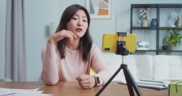Attraktiv ung asiatisk kvinde sidder ved bordet og optager video vlog på smartphone kamera. Smilende kvindelig vlogger gør live stream, konference og tale med seere, tilhængere. Blogging. – Stock-video