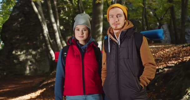 Plano medio de joven hermosa pareja de pie sonriendo en el bosque. Hombre y mujer turista en sombreros de colores mirando a la cámara en el paisaje de bosques. Concepto de personas que viajan en la naturaleza. — Vídeo de stock
