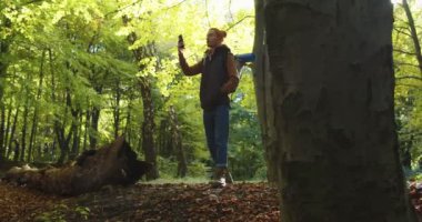 Şapkalı bir gezginin ormanda cep telefonu sinyalini yakalamaya çalışmasının orta planı, telefondan sinyal alamaması. Orman manzarasının arka planında erkek turist. Doğada seyahat eden insanlar kavramı.